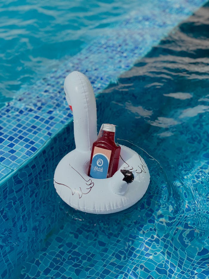 A vibrant red juice bottle floats on a white swan pool float in a blue swimming pool.